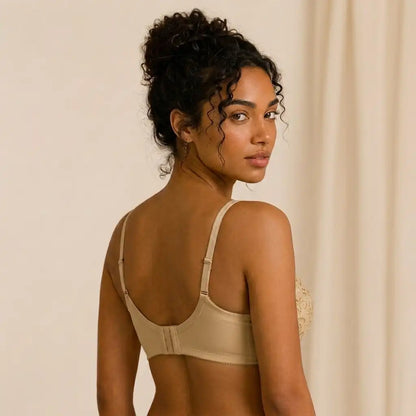 A woman with curly dark hair is shown from the back wearing a beige underwire bra with adjustable shoulder straps, set against a soft beige curtain background.